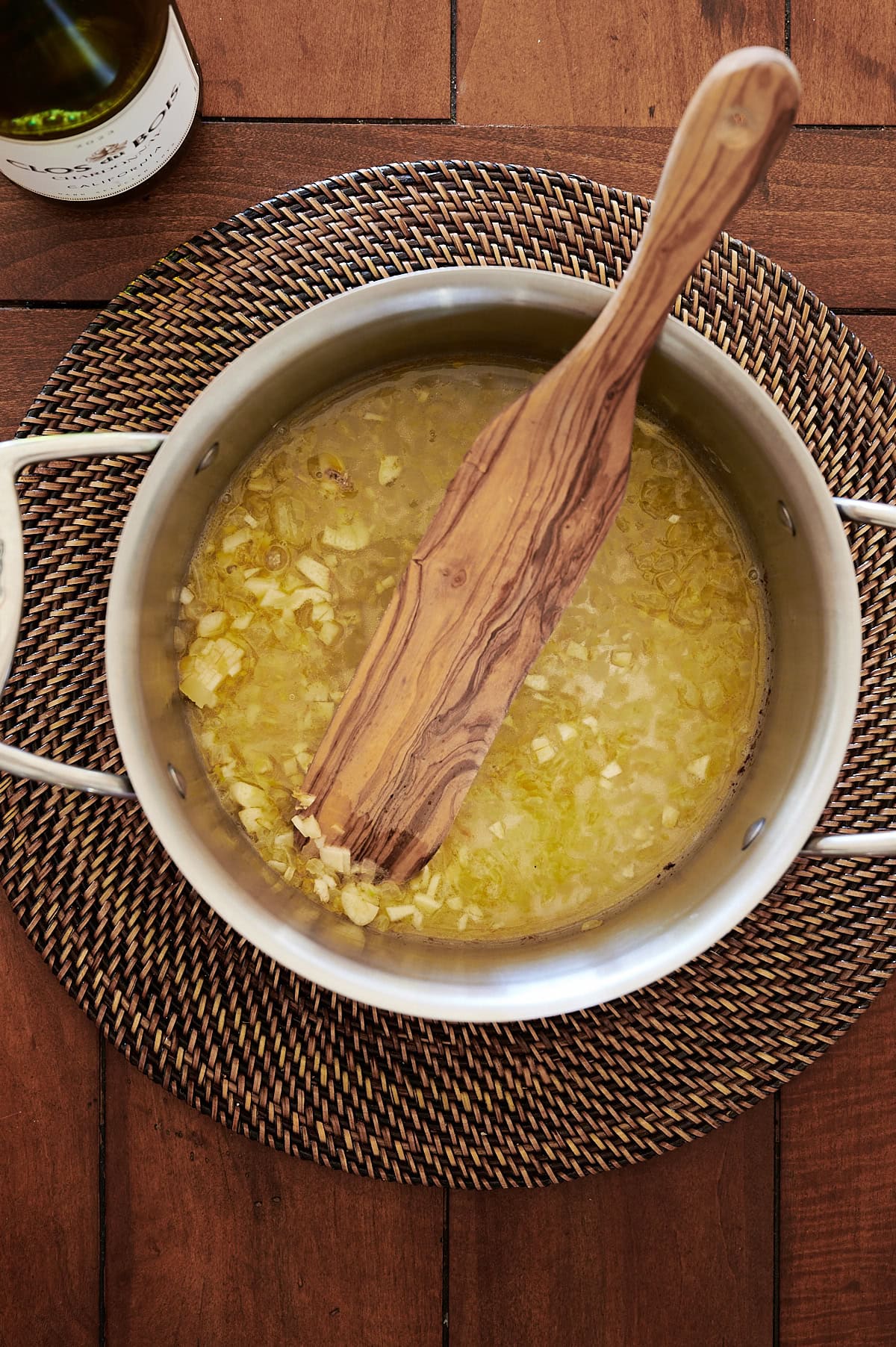 A saucepan with chopped onions cooking in oil, stirred with a wooden spatula, sits on a woven placemat on a wooden table.