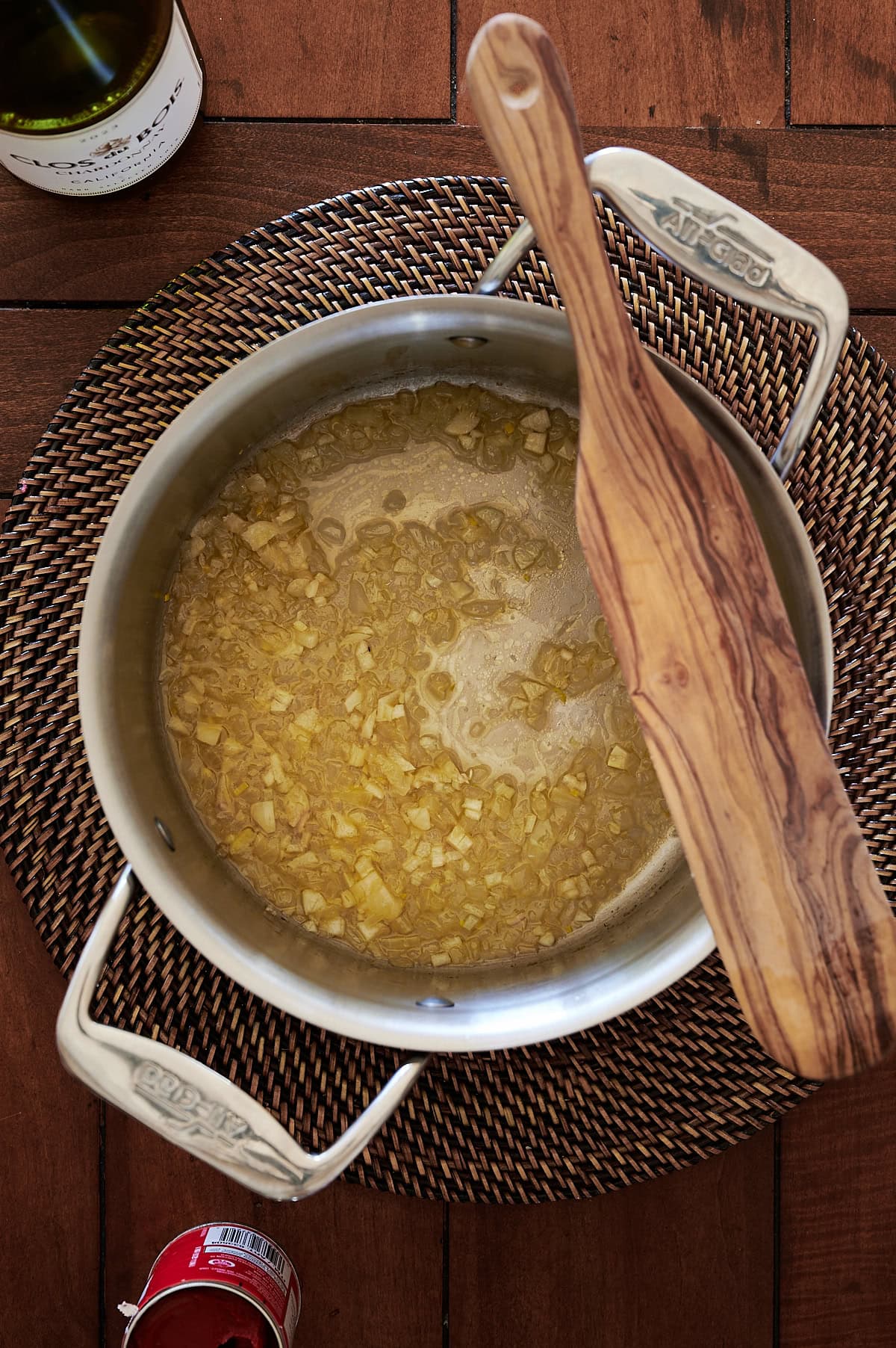 A stainless steel pot with sautéed onions sits on a woven placemat, with a wooden spoon resting on top; wine bottle and canned tomato partially visible.