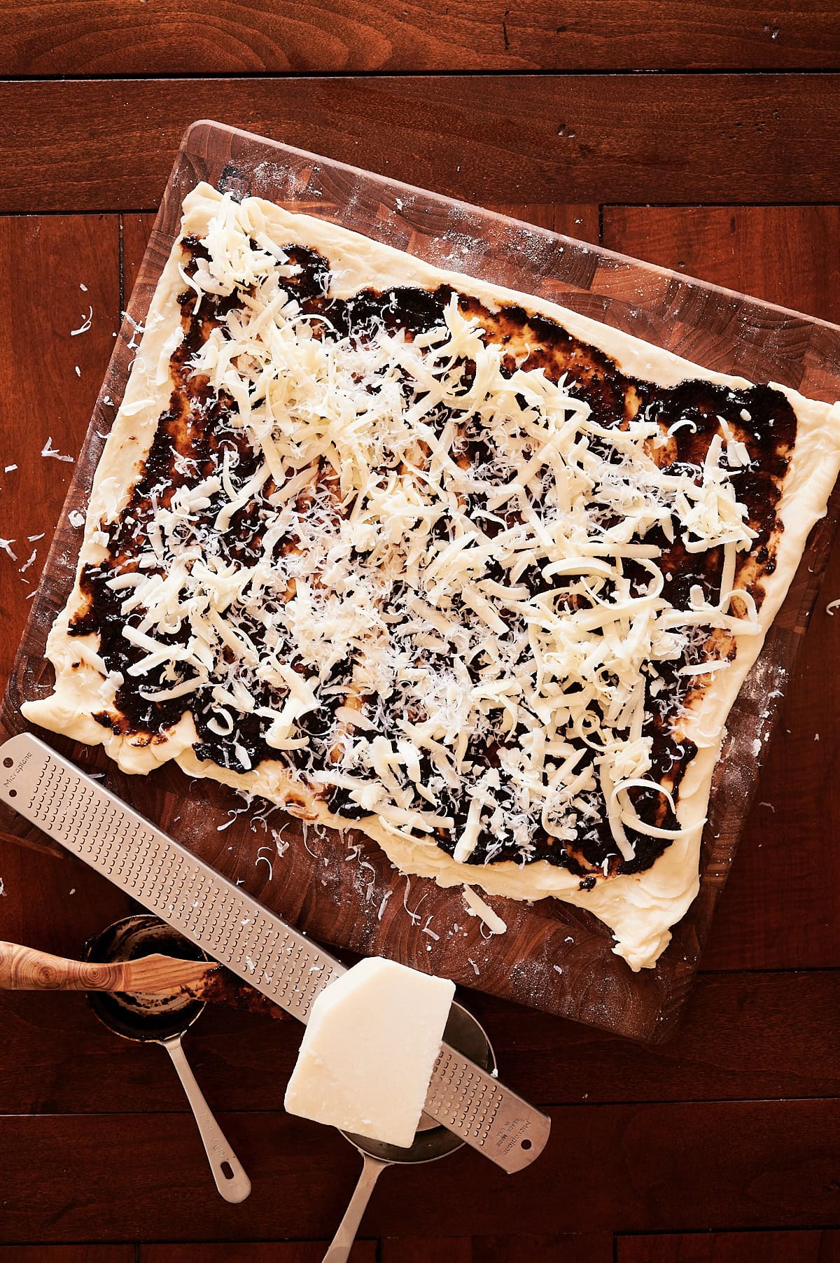 A rolled-out pizza dough topped with a fig jam and shredded cheese sits on a cutting board; cheese block, grater, and small sauce bowl are nearby.