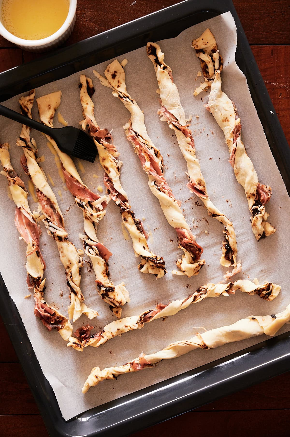 Unbaked twisted pastry cheese straws with fig filling are arranged on a parchment-lined baking tray, with a pastry brush and a cup of egg wash nearby.