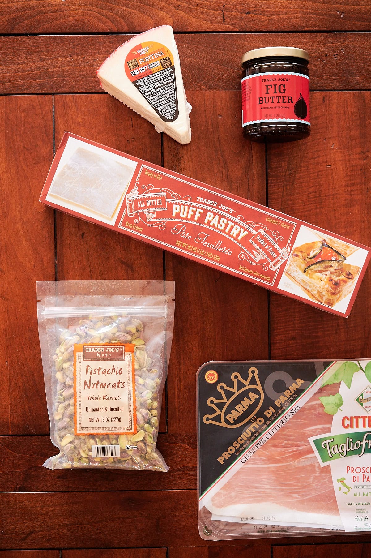 A wedge of cheese, a jar of fig butter, a box of puff pastry, a bag of shelled pistachios, and a package of prosciutto displayed on a wooden surface.