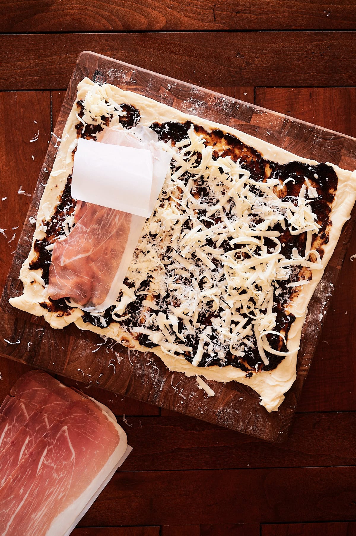 A sheet of dough topped with shredded cheese, fig jam, and slices of prosciutto, partially rolled on a wooden surface.