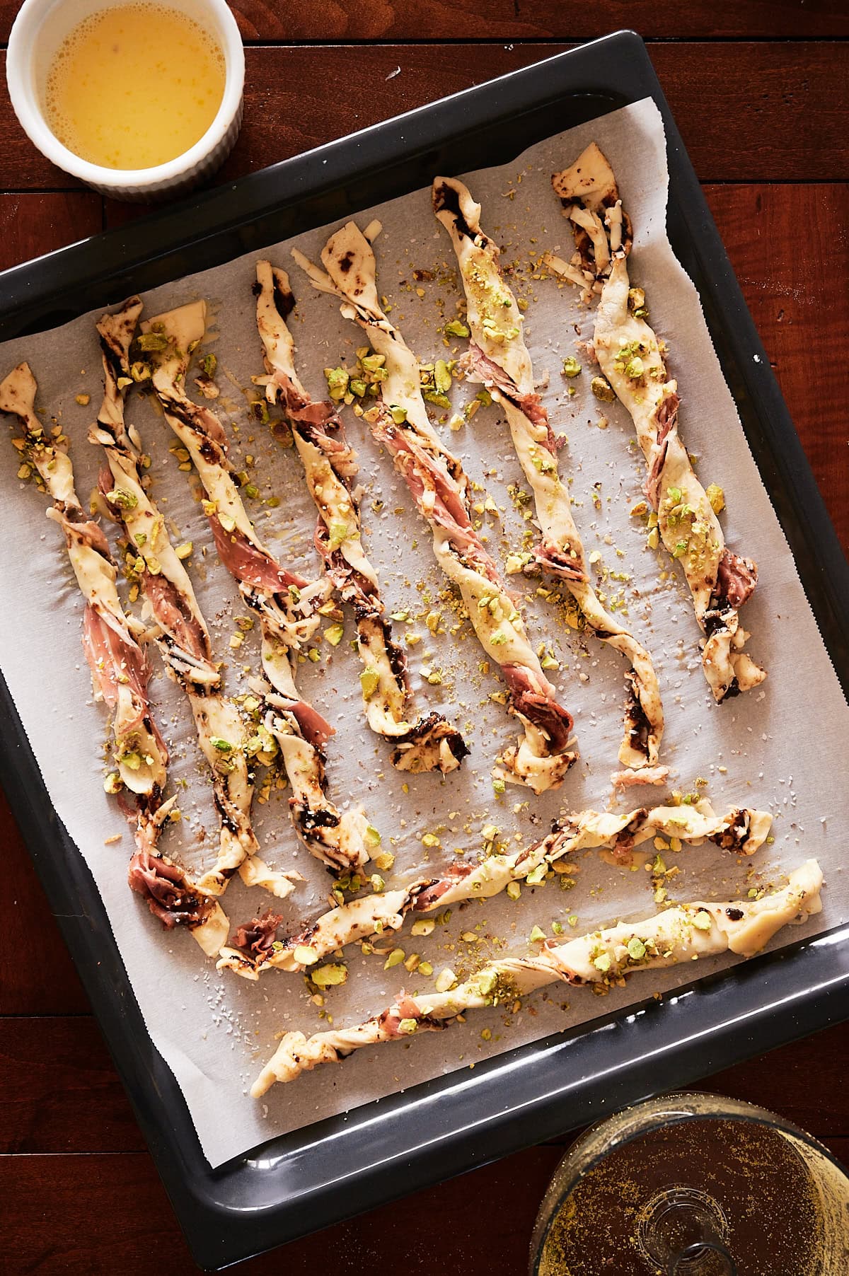 Unbaked pastry cheese straws with chopped pistachios and prosciutto on parchment-lined baking tray, ready for the oven; bowl of egg wash nearby.