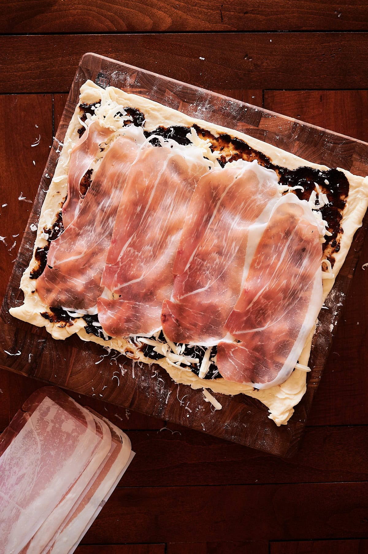 A rectangular pastry topped with grated cheese, fig jam, and several slices of prosciutto on a wooden surface, with a pack of prosciutto nearby.