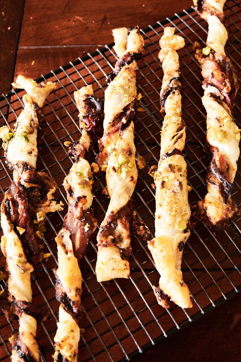 Several twisted puff pastry straws with fig filling rest on a cooling rack atop a wooden surface.