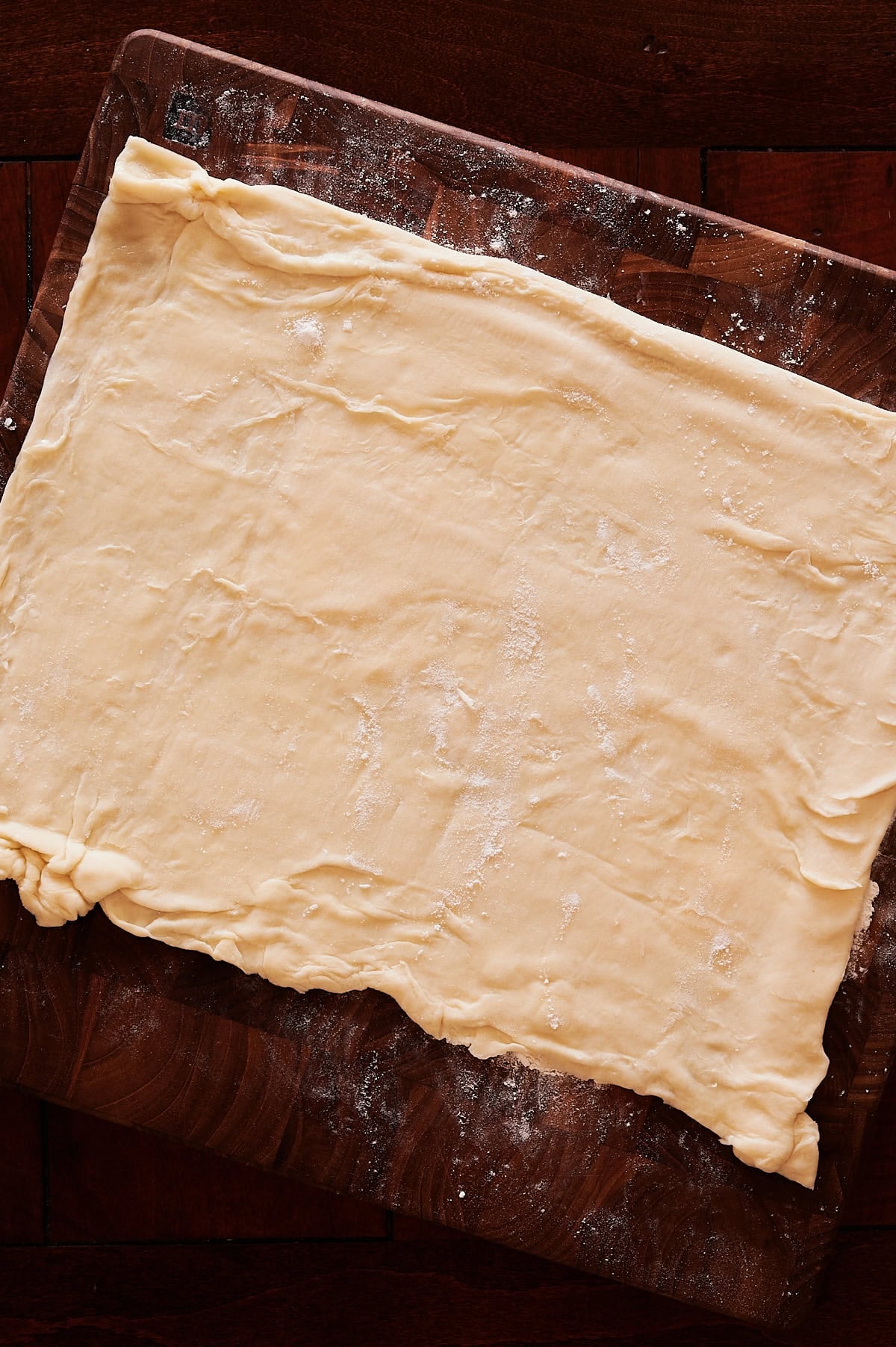A large, uneven rectangle of rolled-out puff pastry rests on a floured wooden surface.