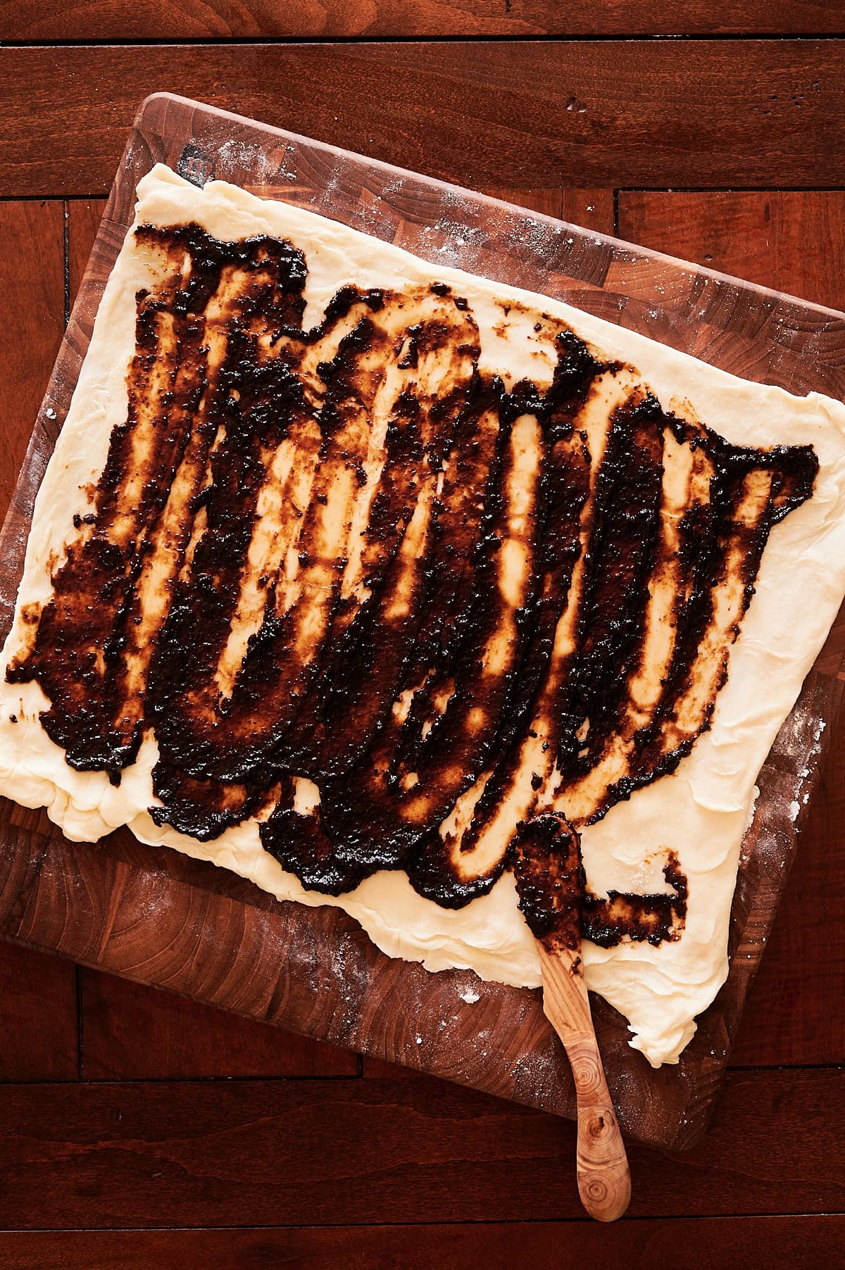 Rolled-out dough on a floured wooden board spread with a fig jam; a wooden spatula rests on the corner.