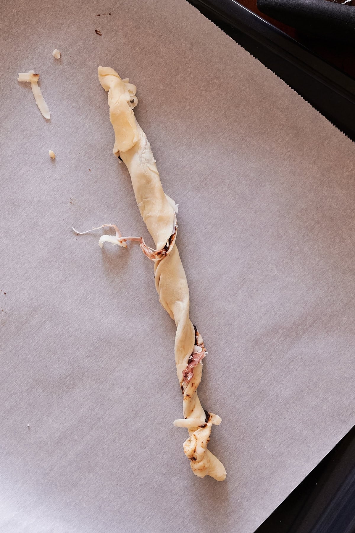 Unbaked twisted puff pastry cheese straw with visible cheese and prosciutto placed on a sheet of parchment paper on a baking tray.