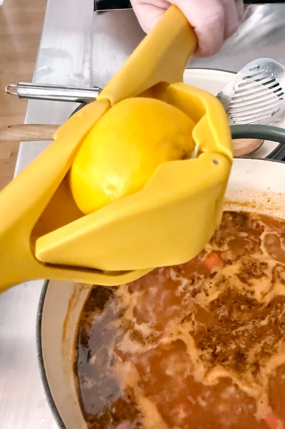 A hand uses a yellow citrus squeezer to press a lemon over a pot of simmering pot of soup on a stove.