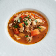 A white bowl filled with chicken soup containing diced carrots, herbs, farro, and chopped chicken, garnished with fresh parsley.