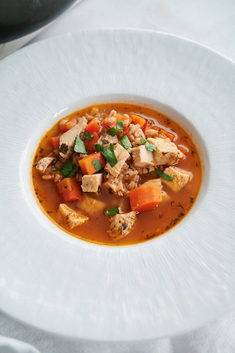 A white bowl filled with chicken soup containing diced carrots, herbs, farro, and chopped chicken, garnished with fresh parsley.