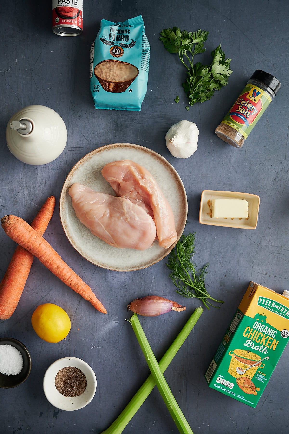 Raw chicken breasts on a plate surrounded by carrots, celery, butter, garlic, shallot, parsley, dill, lemon, farro, chicken broth, tomato paste, seasoning, salt, pepper, and olive oil.