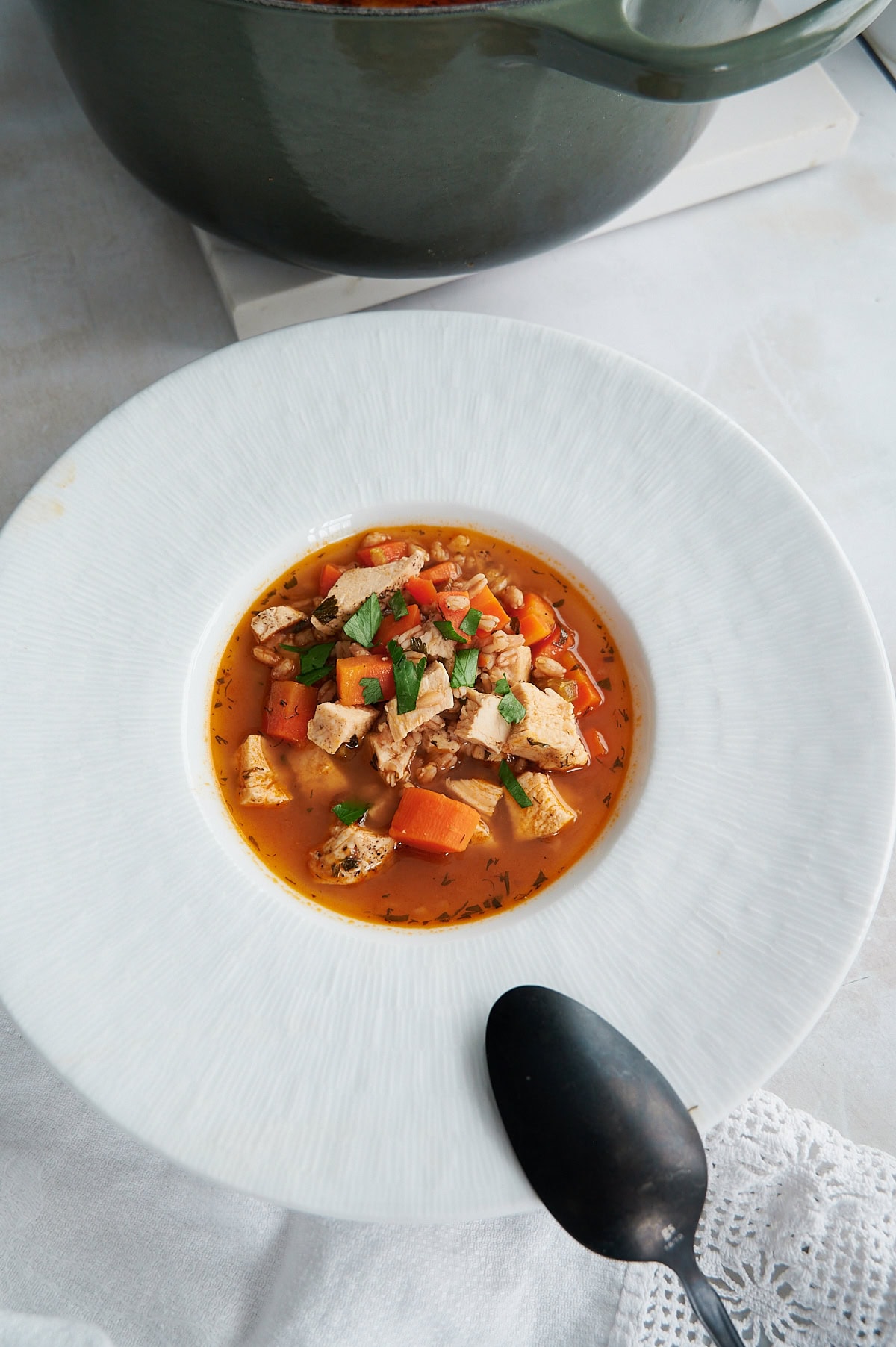 A white bowl filled with farro, vegetable and chicken soup sits on a table beside a black spoon and a green pot.