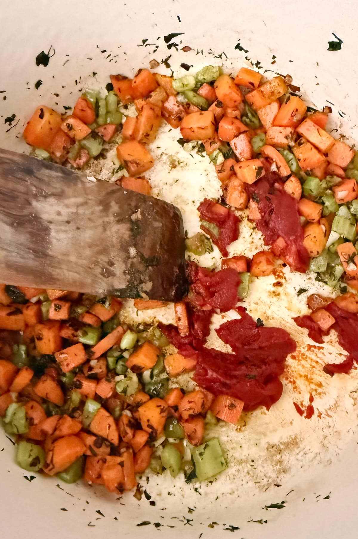 Diced carrots and celery are sautéed with tomato paste and herbs in a pan, being stirred with a wooden spatula.