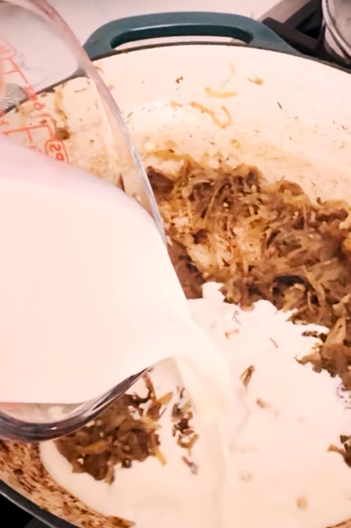 Cream is being poured from a measuring cup into a pot containing caramelized onions.