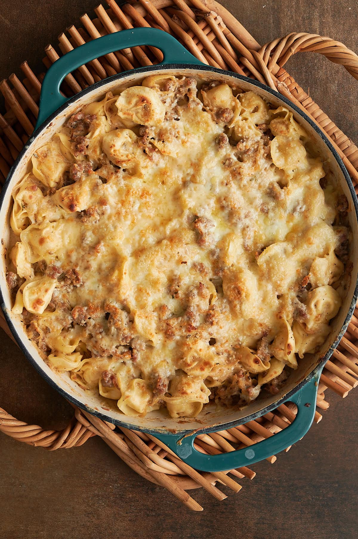 A baked tortellini casserole with melted cheese and ground sausage in a round blue baking dish on a woven placemat.