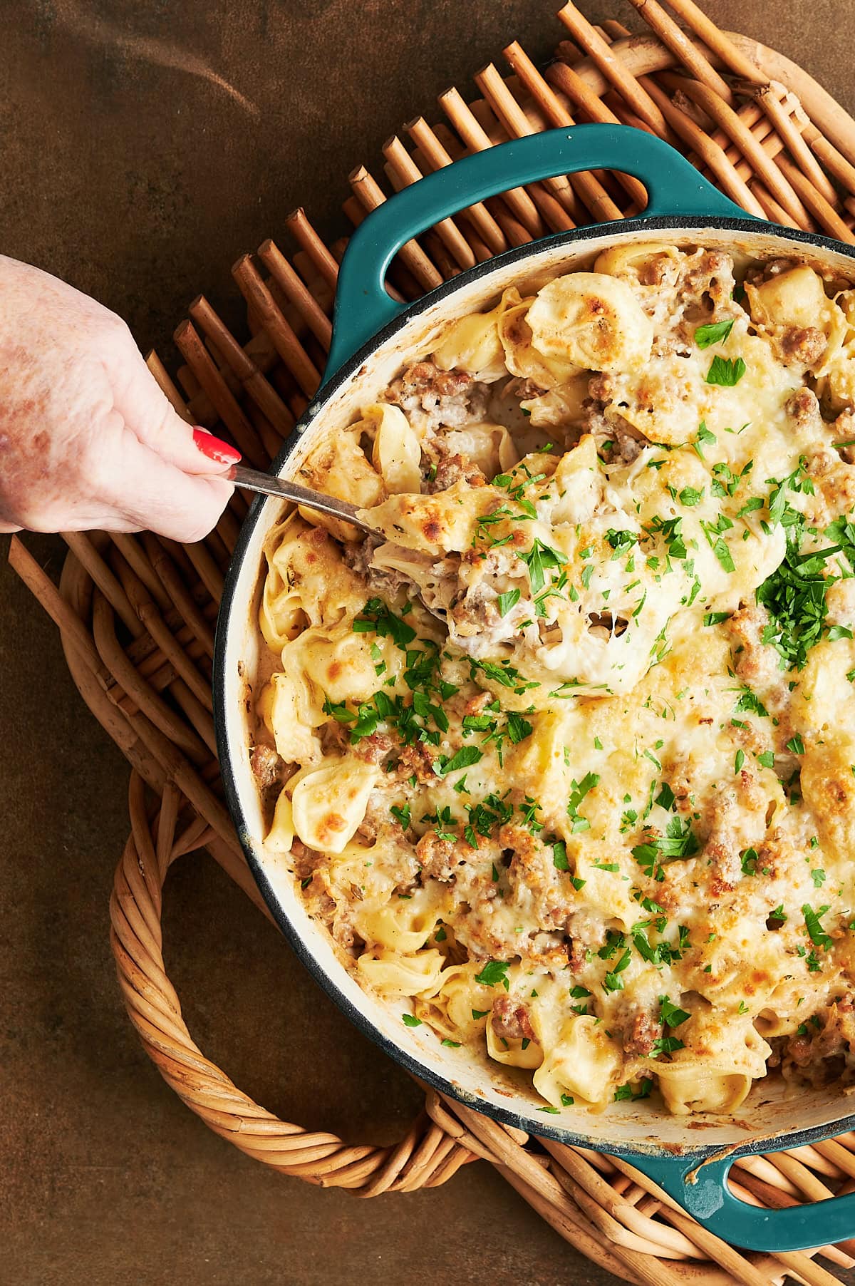 A hand serves cheesy tortellini pasta with ground sausage and parsley from a blue casserole dish on a woven placemat.