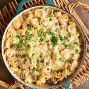 A baked tortellini casserole with ground sausage, cheese, and parsley in a blue skillet on a woven trivet.