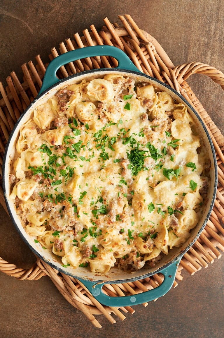 A baked tortellini casserole with ground sausage, cheese, and parsley in a blue skillet on a woven trivet.