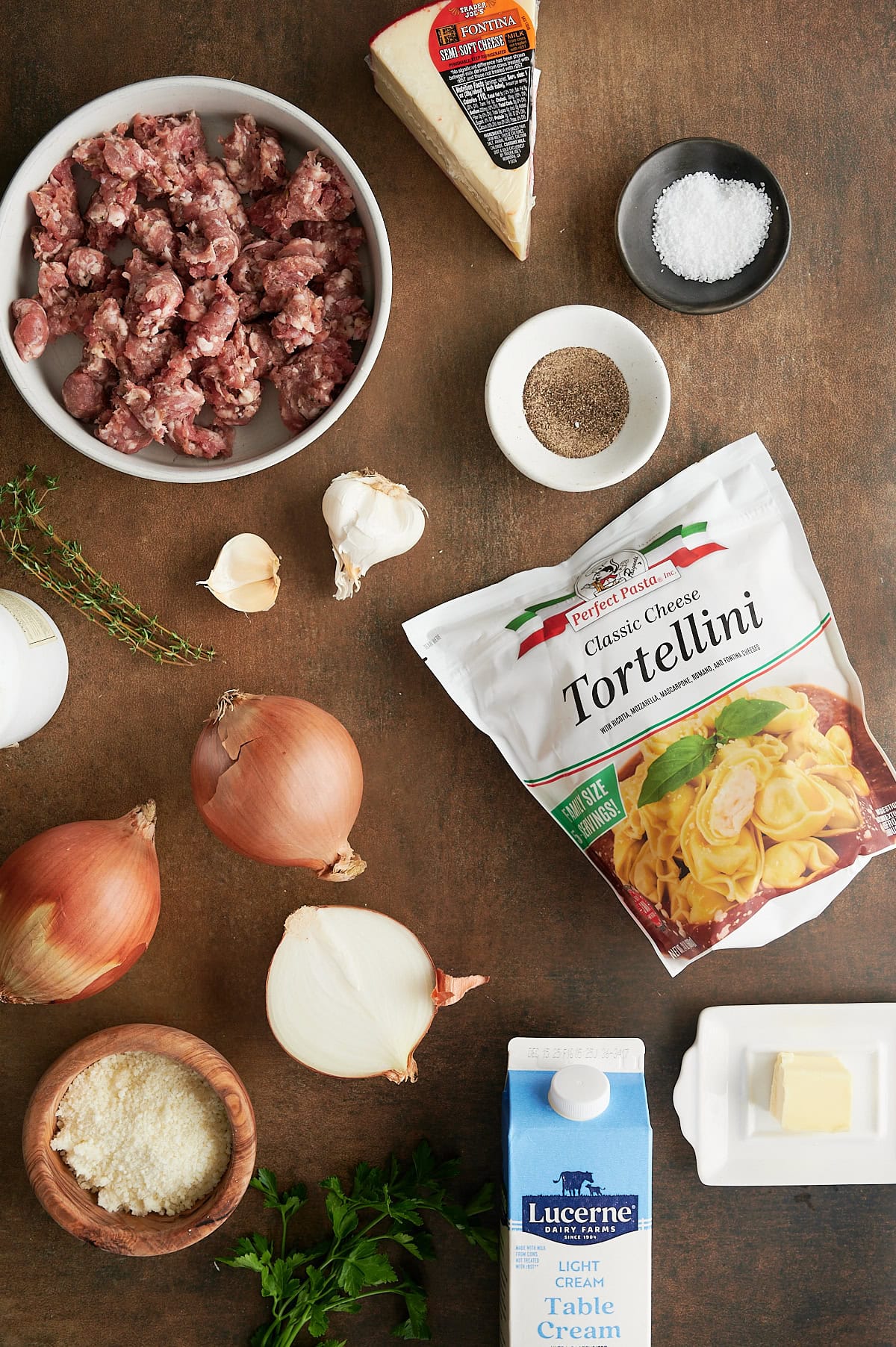 Ingredients for a tortellini pasta baked dish including ground sausage, Parmesan, salt, pepper, garlic, tortellini, onions, cream, butter, parsley, and thyme arranged on a brown surface.