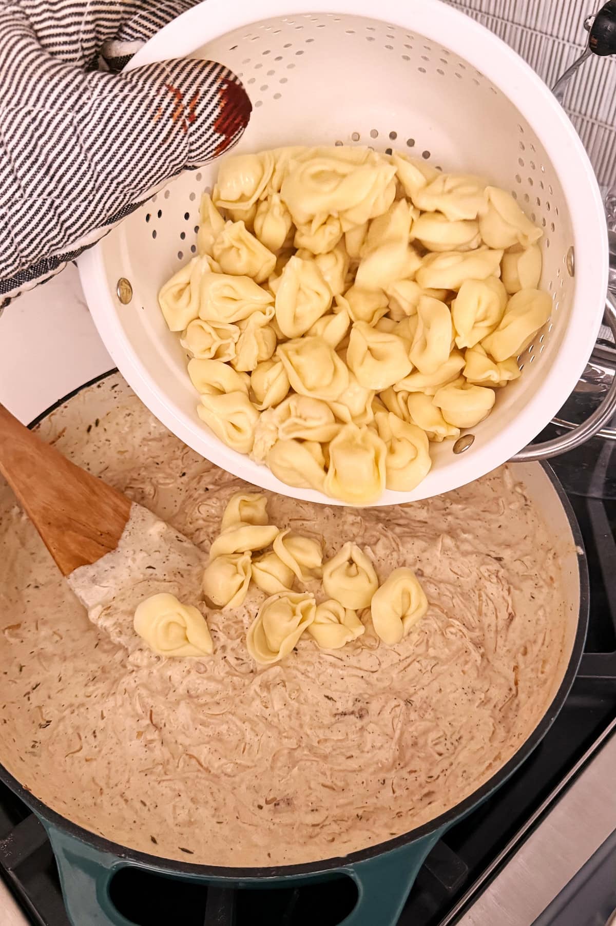 A hand pours cheese tortellini from a white colander into a pot of creamy sauce on a stovetop while stirring with a wooden spatula.