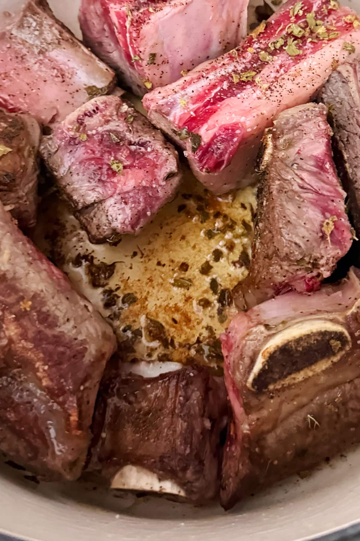 Several pieces of seasoned beef short ribs are being browned in a pot, with visible searing and rendered fat on the surface.