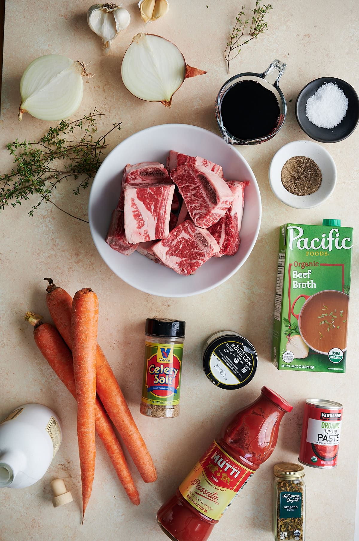 A flat lay of ingredients for a braised short ribs, including raw short ribs, carrots, onion, garlic, thyme, broth, tomato sauce, tomato paste, wine, spices, salt, and pepper.