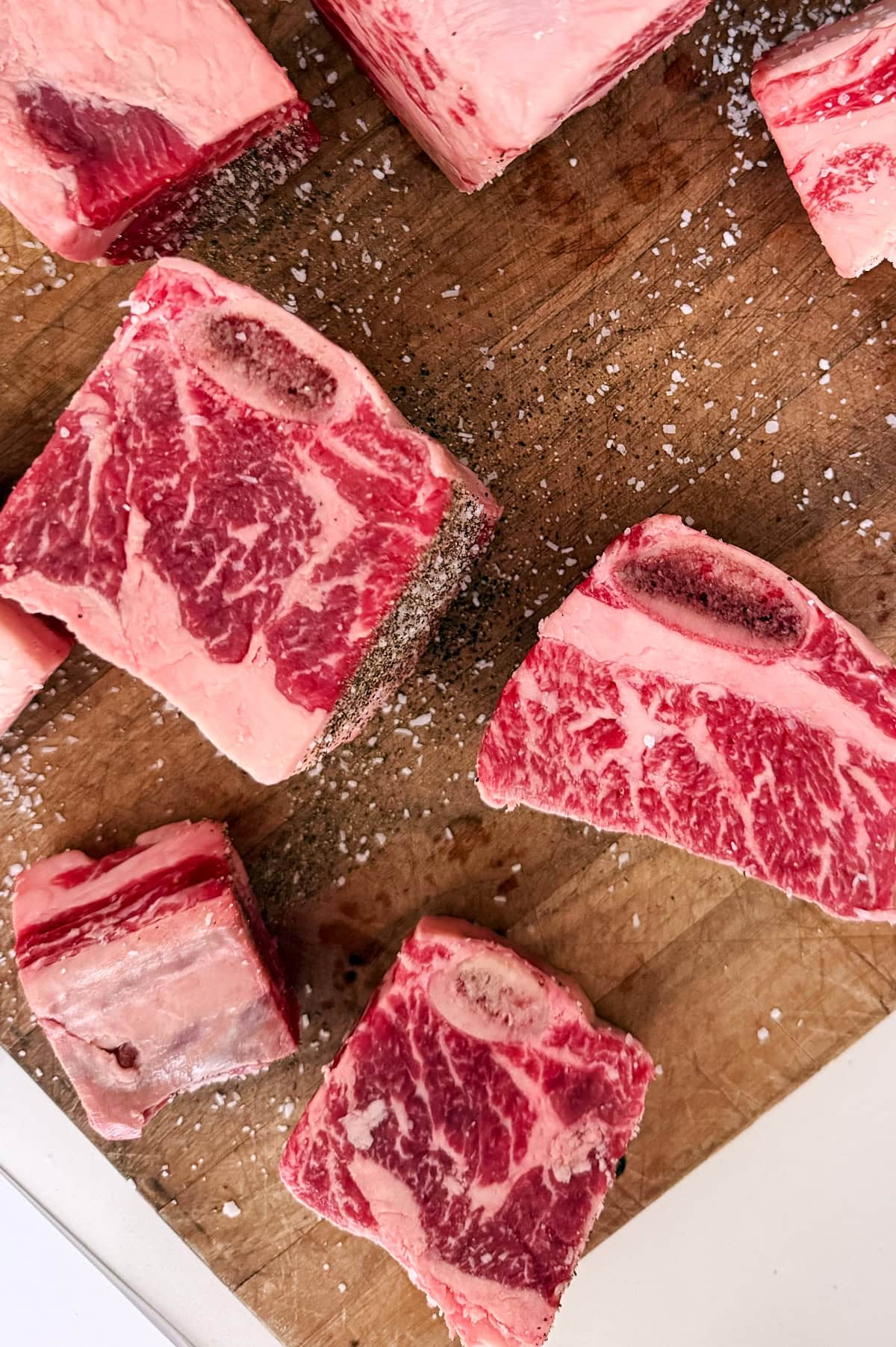 Several thick, raw beef short rib pieces with marbled fat are arranged on a wooden cutting board sprinkled with coarse salt.