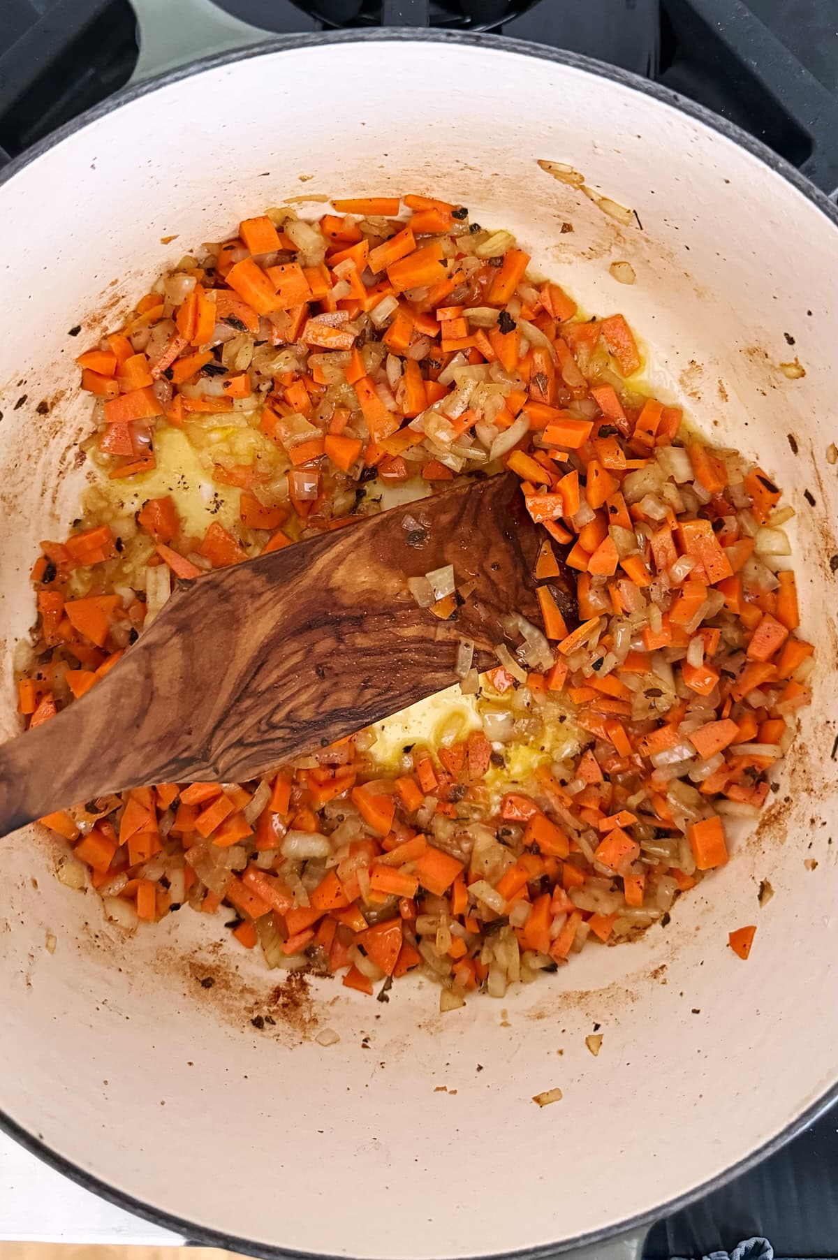Chopped carrots and onions being sautéed in oil in a white pot, stirred with a wooden spatula.