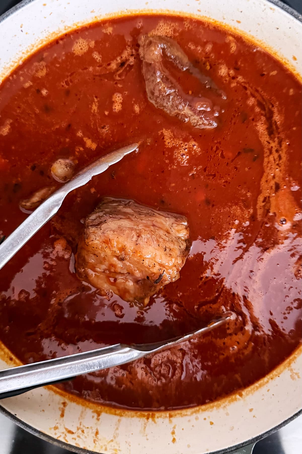 A pot of red tomato-based sauce with a beef short rib, with metal tongs resting inside.