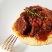 A serving of braised short ribs with carrots and sauce on polenta garnished with a sprig of thyme, on a white plate next to a fork.