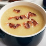 A bowl of creamy soup topped with crispy bacon pieces, with a plate of extra bacon in the background.