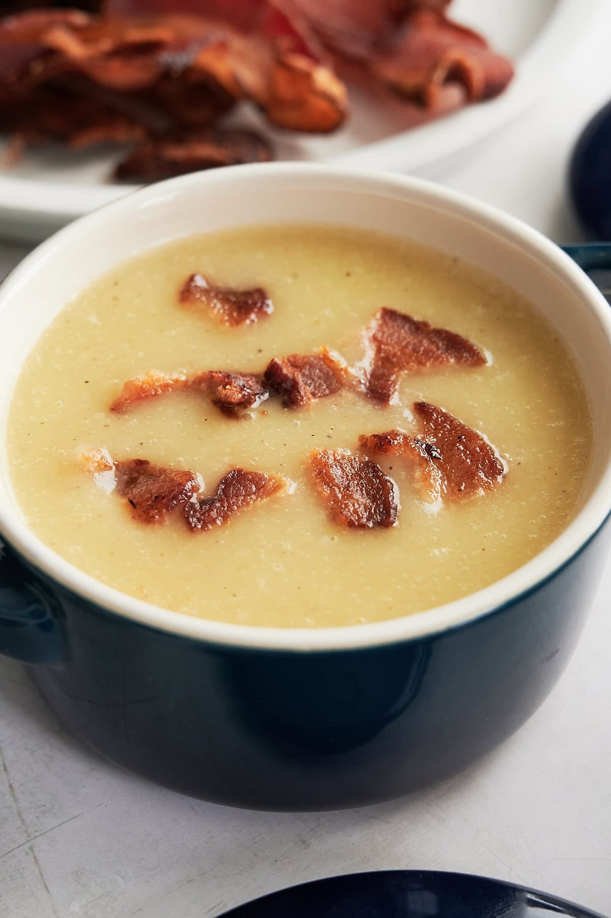 A bowl of creamy soup topped with crispy bacon pieces, with a plate of extra bacon in the background.