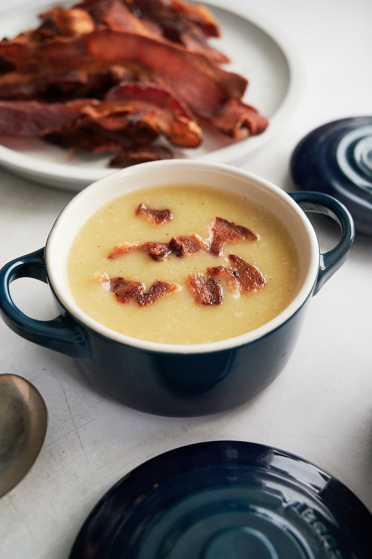 A bowl of creamy leek and potato soup topped with pieces of crispy bacon sits on a table, with a plate of additional bacon in the background.