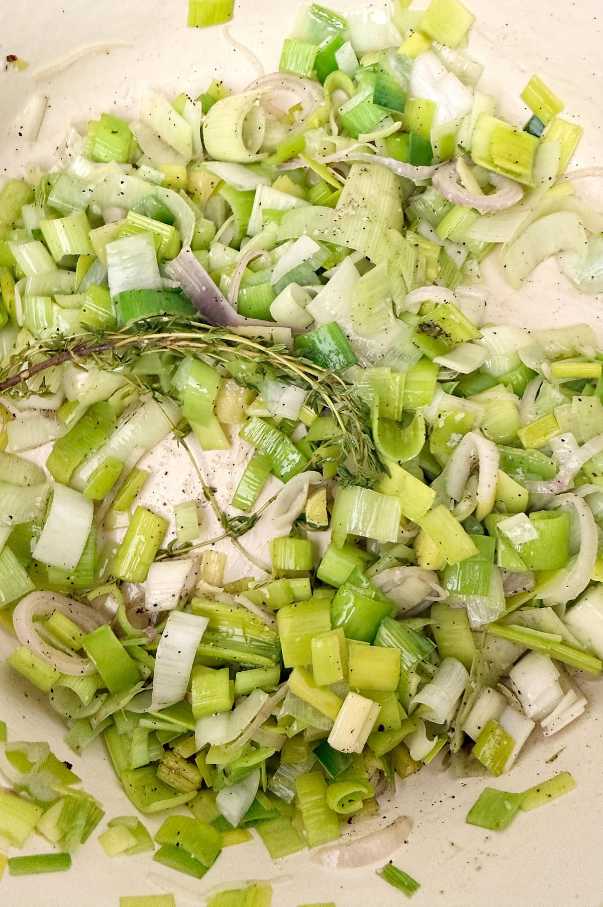 Chopped leeks and fresh thyme sautéing in a pan with visible black pepper and oil.