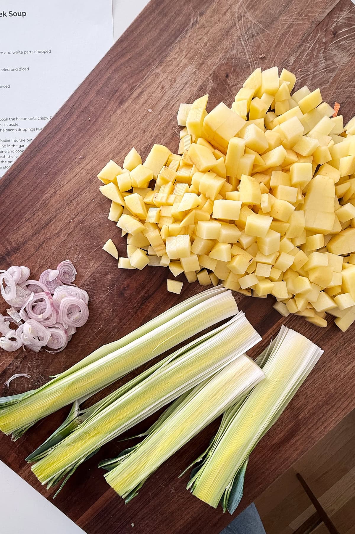 Chopped potatoes, sliced shallots, and halved leeks are arranged on a wooden cutting board, with a printed recipe partially visible in the corner.