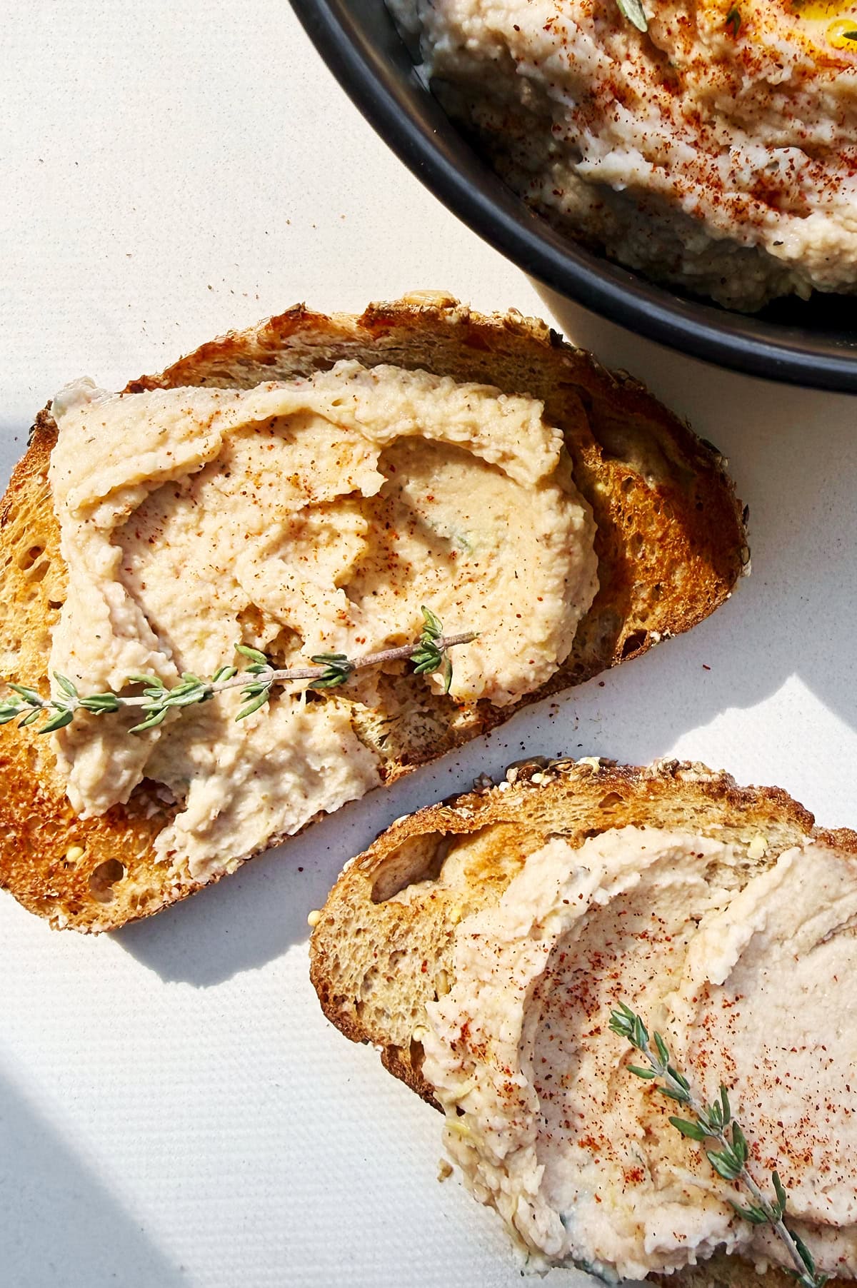 Two slices of bread topped with a creamy cannellini spread, garnished with fresh thyme, next to a bowl containing more of the bean dip on a white surface.
