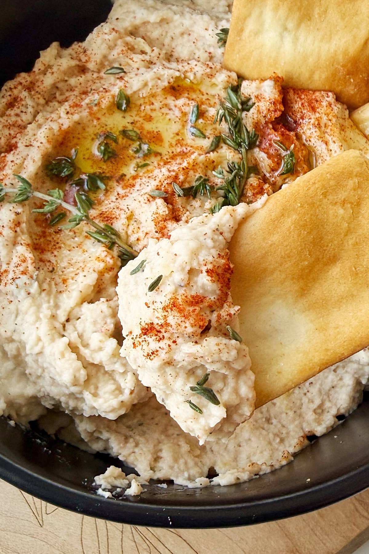 Bowl of cannellini bean dip topped with olive oil, paprika, and herbs, served with pieces of pita chips.