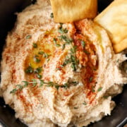 A black bowl of white bean dip garnished with olive oil, paprika, fresh herbs, and served with two pieces of pita bread.