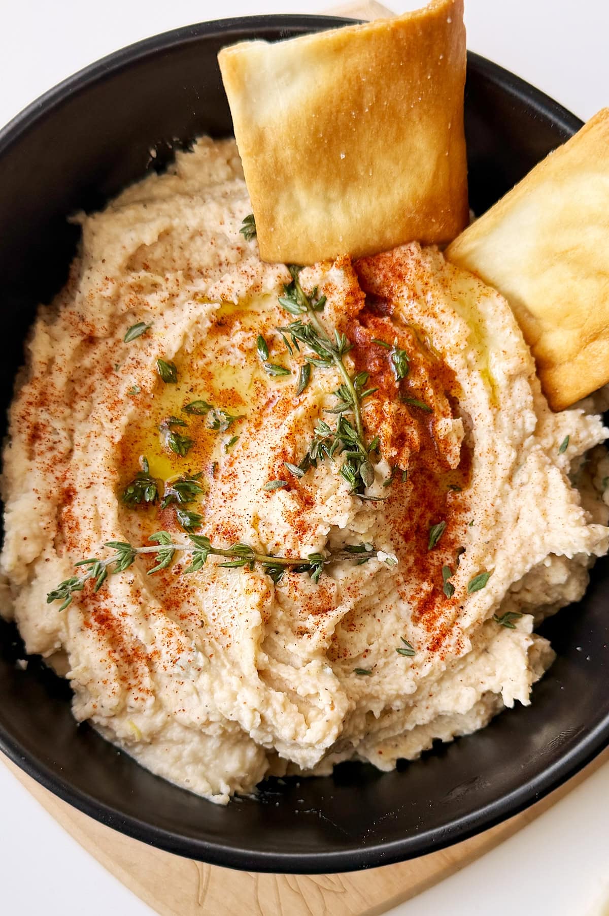 A black bowl of white bean dip garnished with olive oil, paprika, fresh herbs, and served with two pieces of pita bread.
