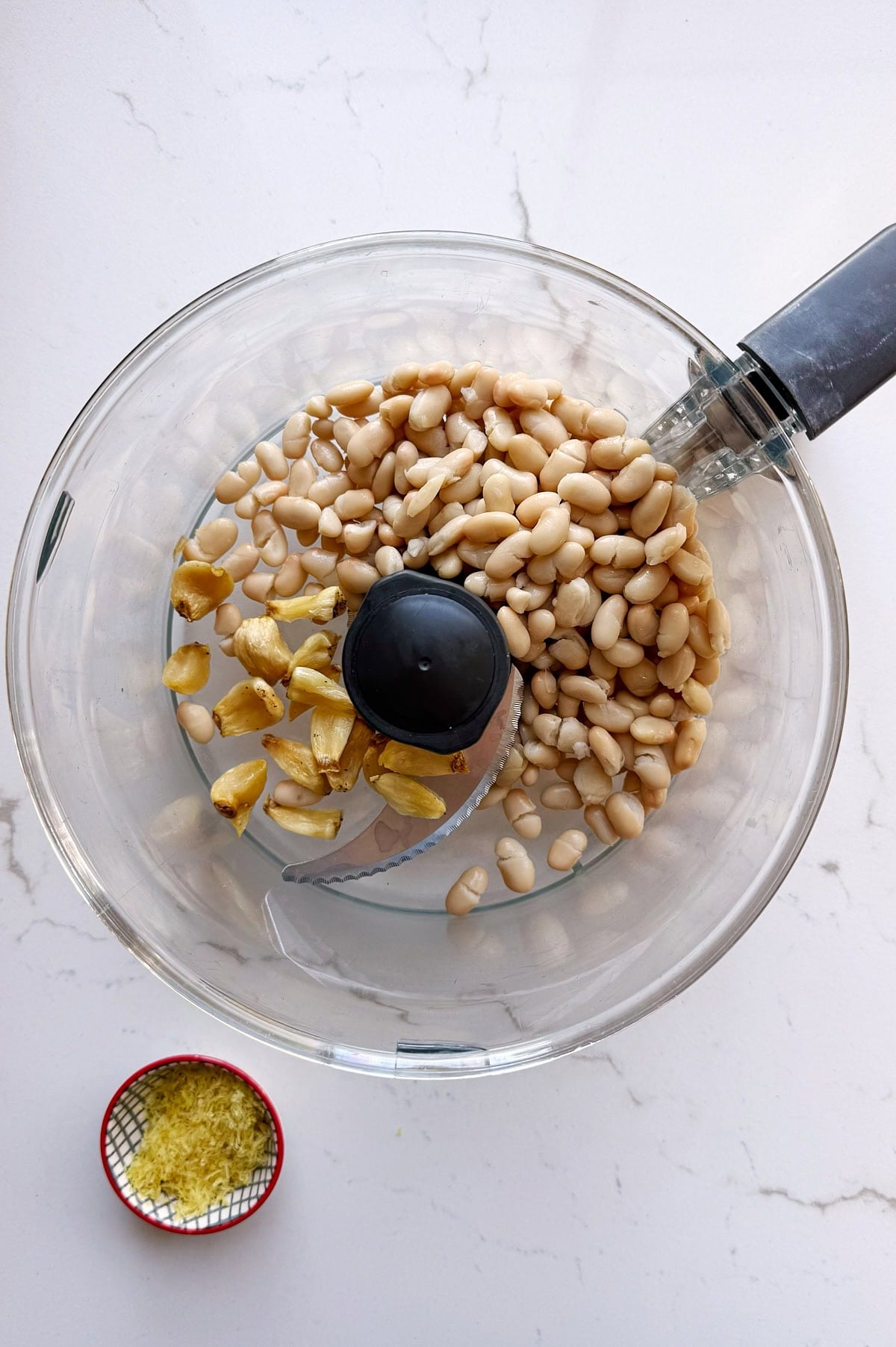 A food processor bowl with white beans and peeled garlic cloves inside, next to a small dish containing lemon zest, on a white countertop.