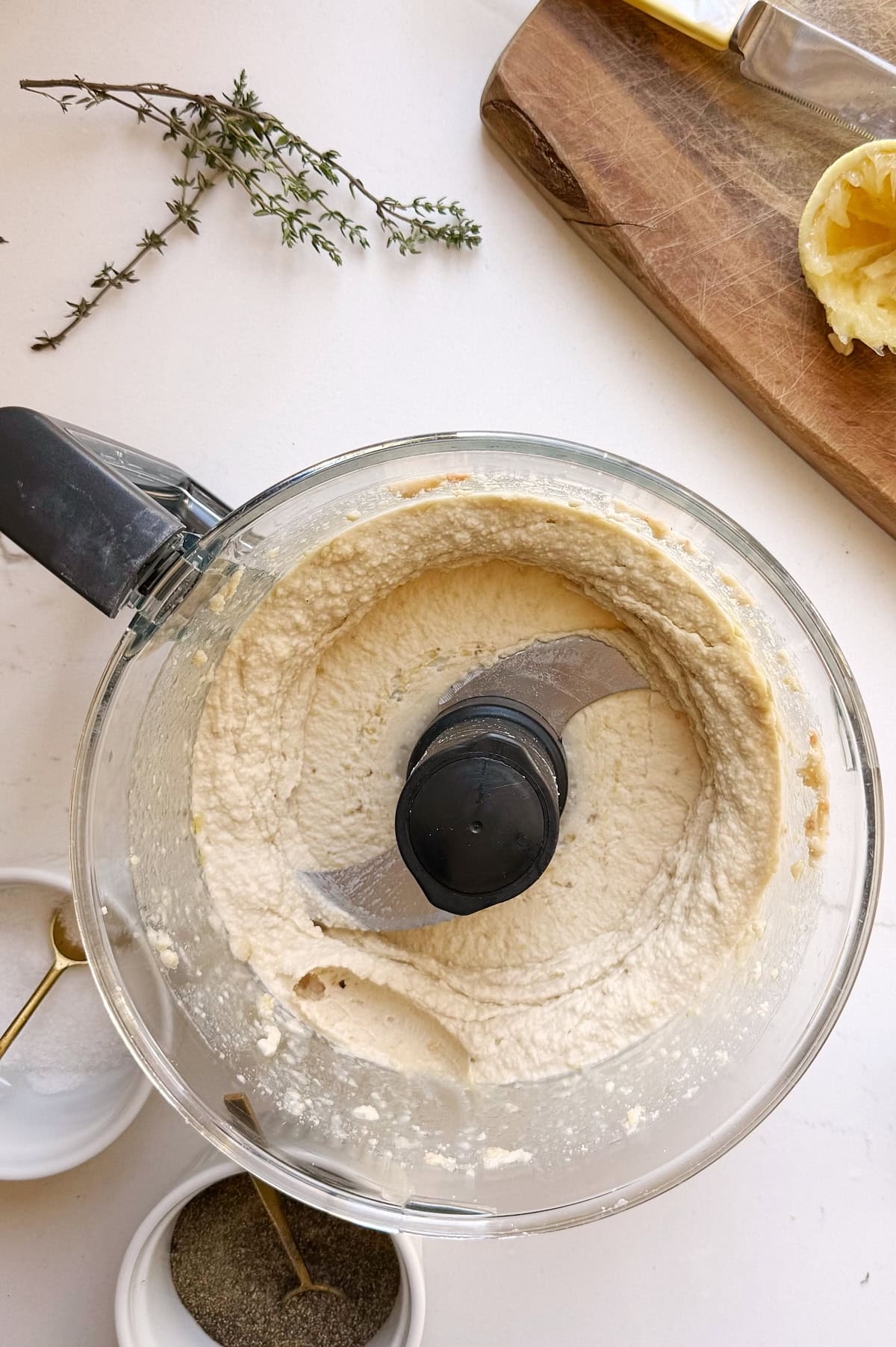 A food processor containing a creamy white bean dip sits on a white countertop near a cutting board, a squeezed lemon half, fresh herbs, salt, and pepper.