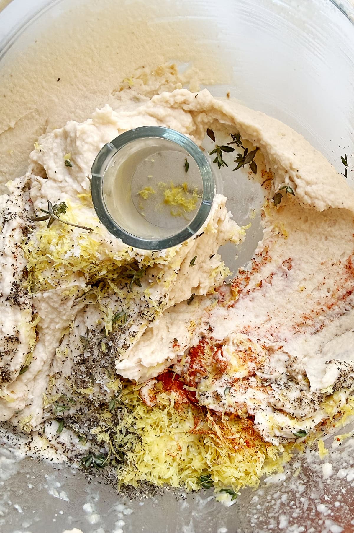 Cannellini bean mixture in a food processor bowl with lemon zest, black pepper, paprika, and fresh herbs on top.