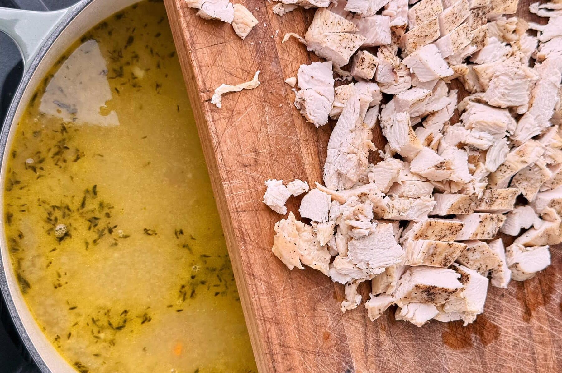 Chopped cooked chicken on a cutting board is about to be added to a pot of soup with broth and herbs.