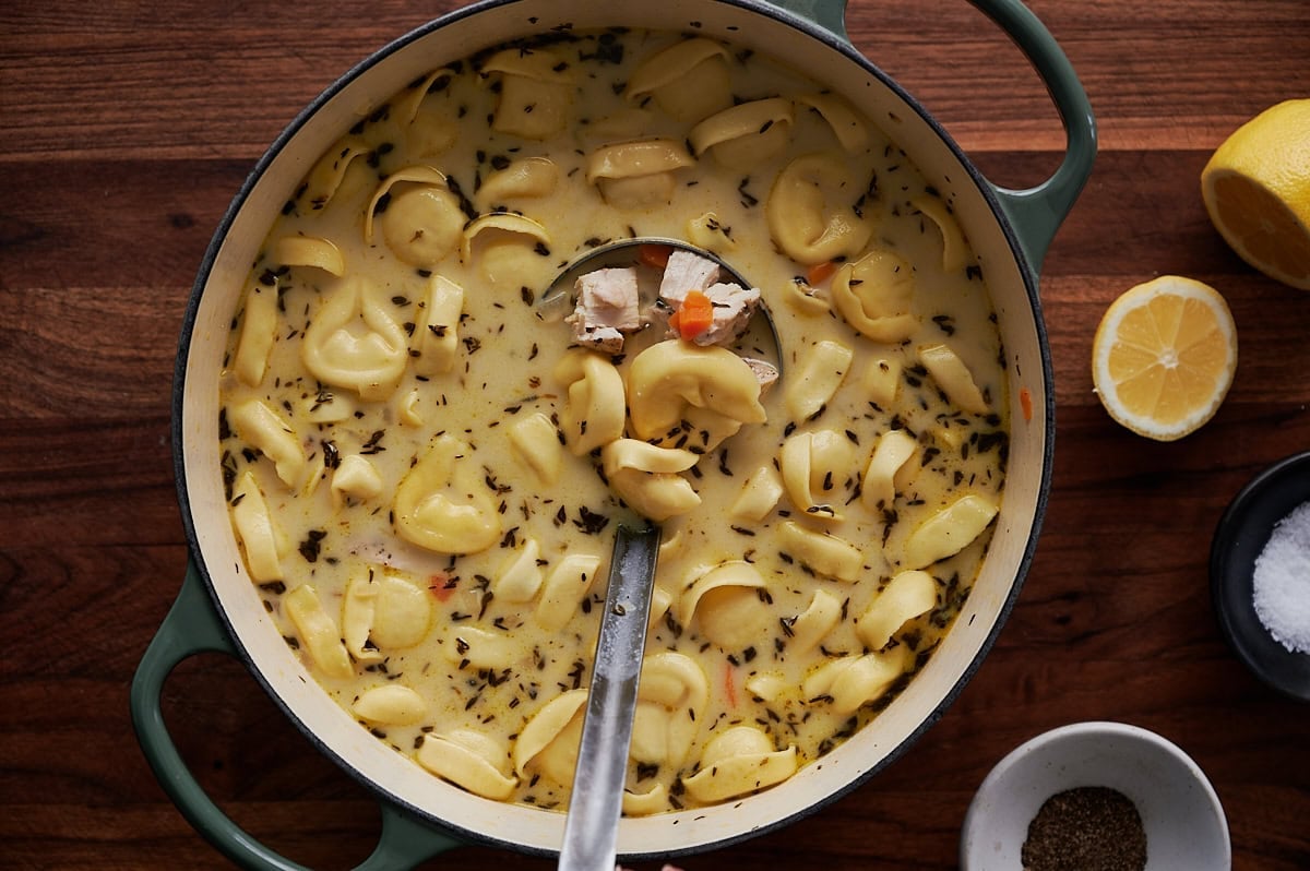 A pot of creamy tortellini soup with herbs, chicken pieces, and vegetables, shown on a wooden table with lemon halves and small dishes of salt and pepper nearby.