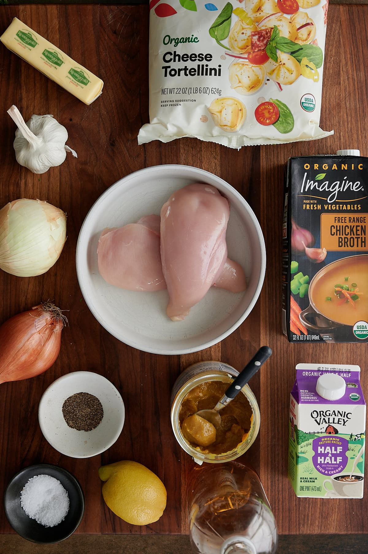 Top-down view of ingredients on a wooden surface, including chicken breasts, cheese tortellini, chicken broth, half & half, garlic, onion, shallot, lemon, butter, spices, and a jar of soup base.