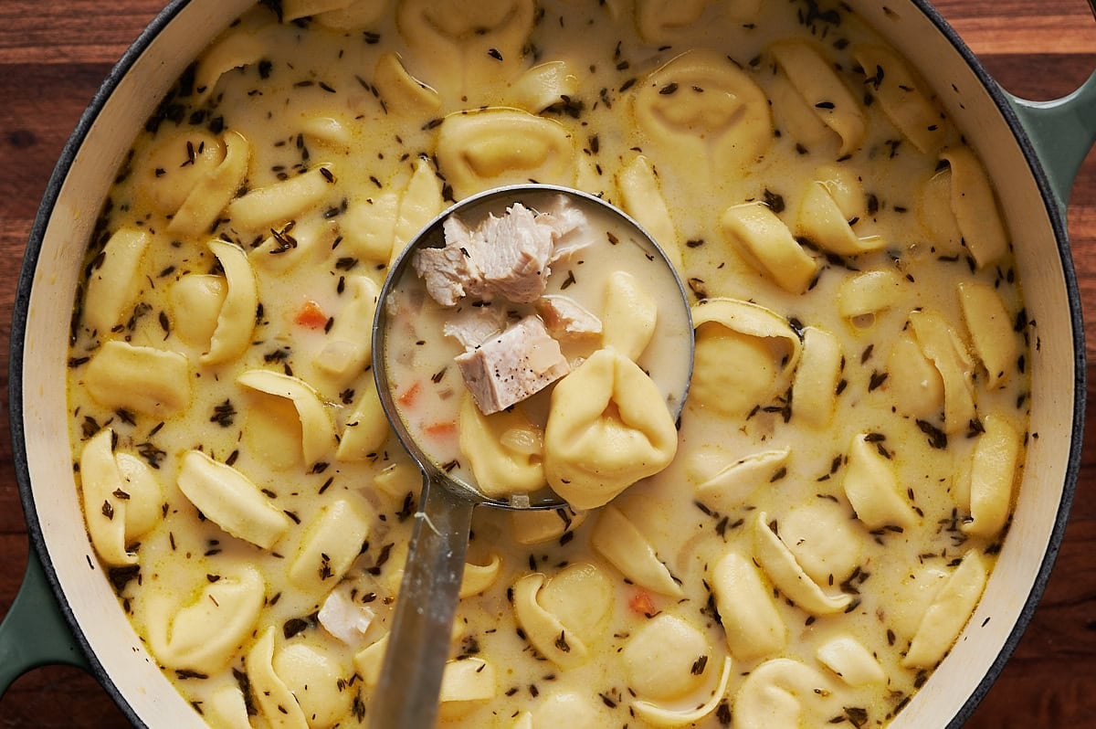 A ladle serving chicken and cheese tortellini soup from a pot, showing pieces of chicken, tortellini pasta, and creamy broth with herbs.