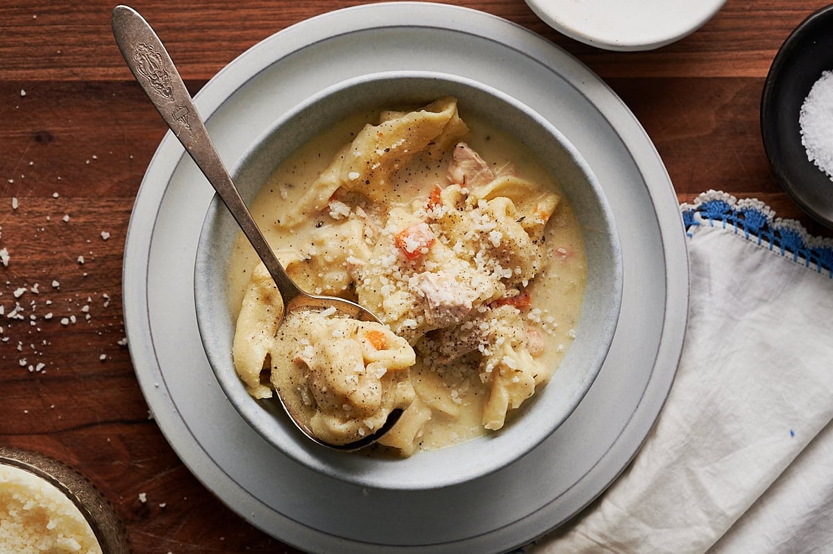 A bowl of creamy chicken and tortellini soup topped with grated cheese, served with a spoon on a wooden table.