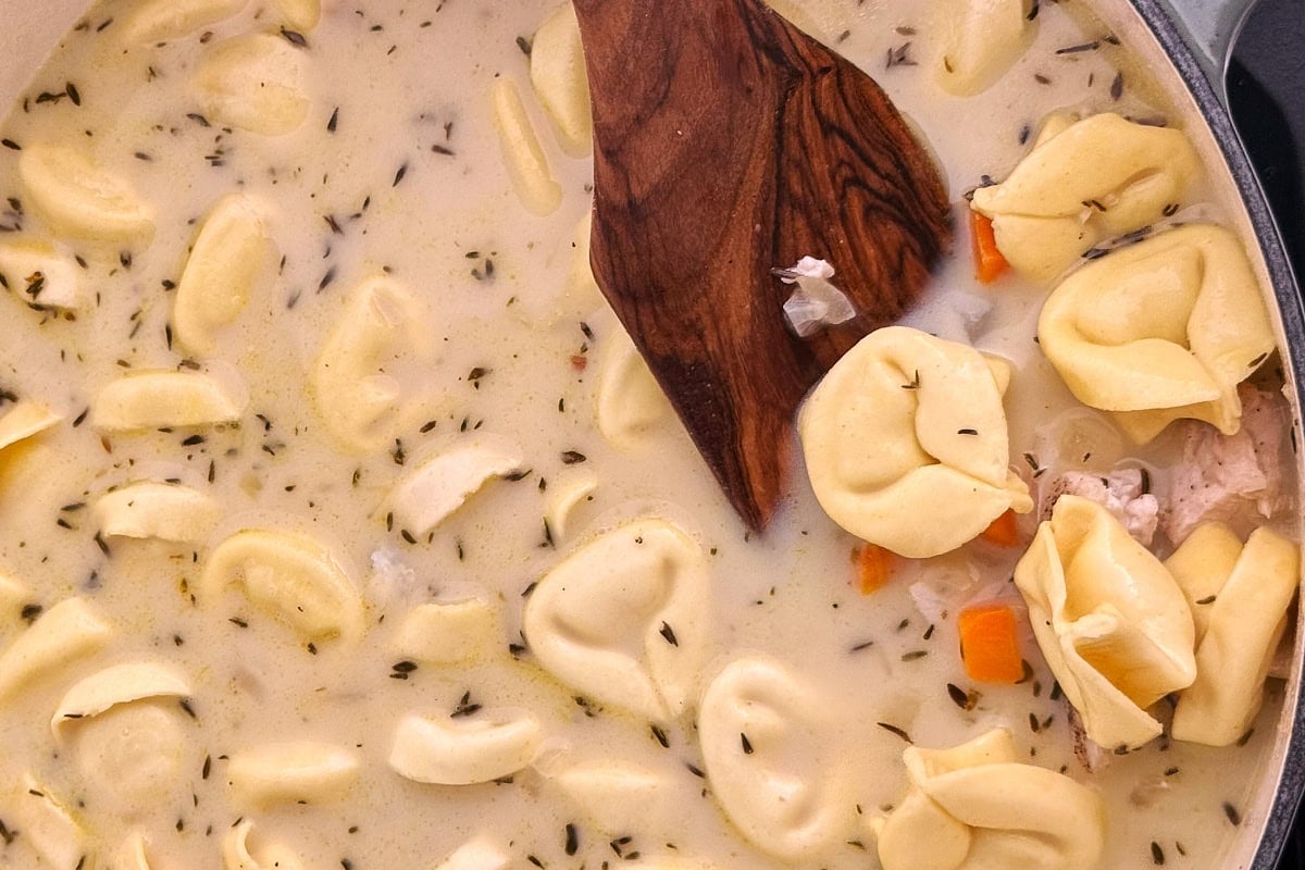 A pot of creamy soup with tortellini pasta, herbs, carrot pieces, and a wooden spoon stirring the mixture.