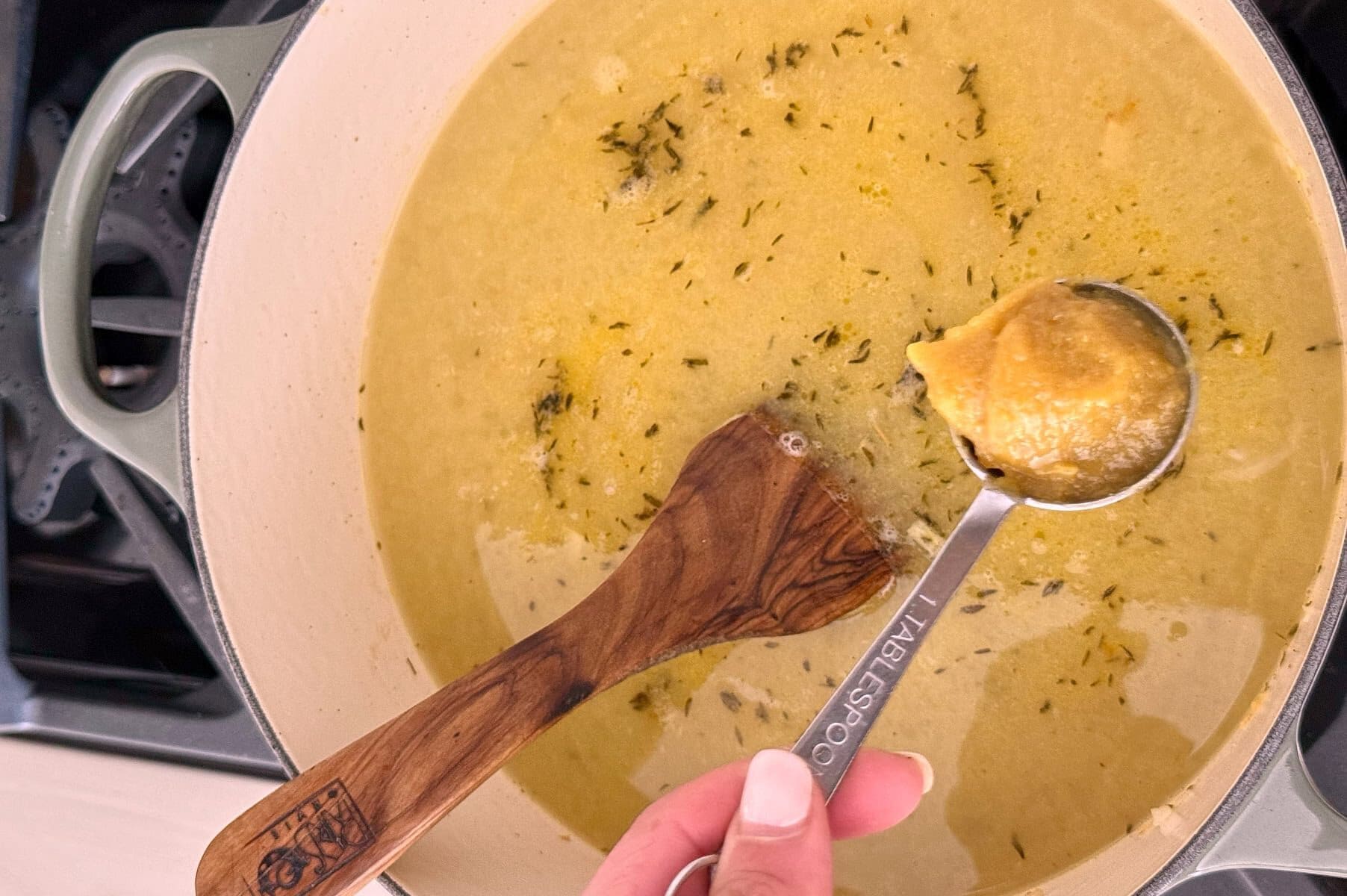 A hand holds a measuring spoon of soup base over a pot of simmering broth with herbs, next to a wooden spoon on a stove.